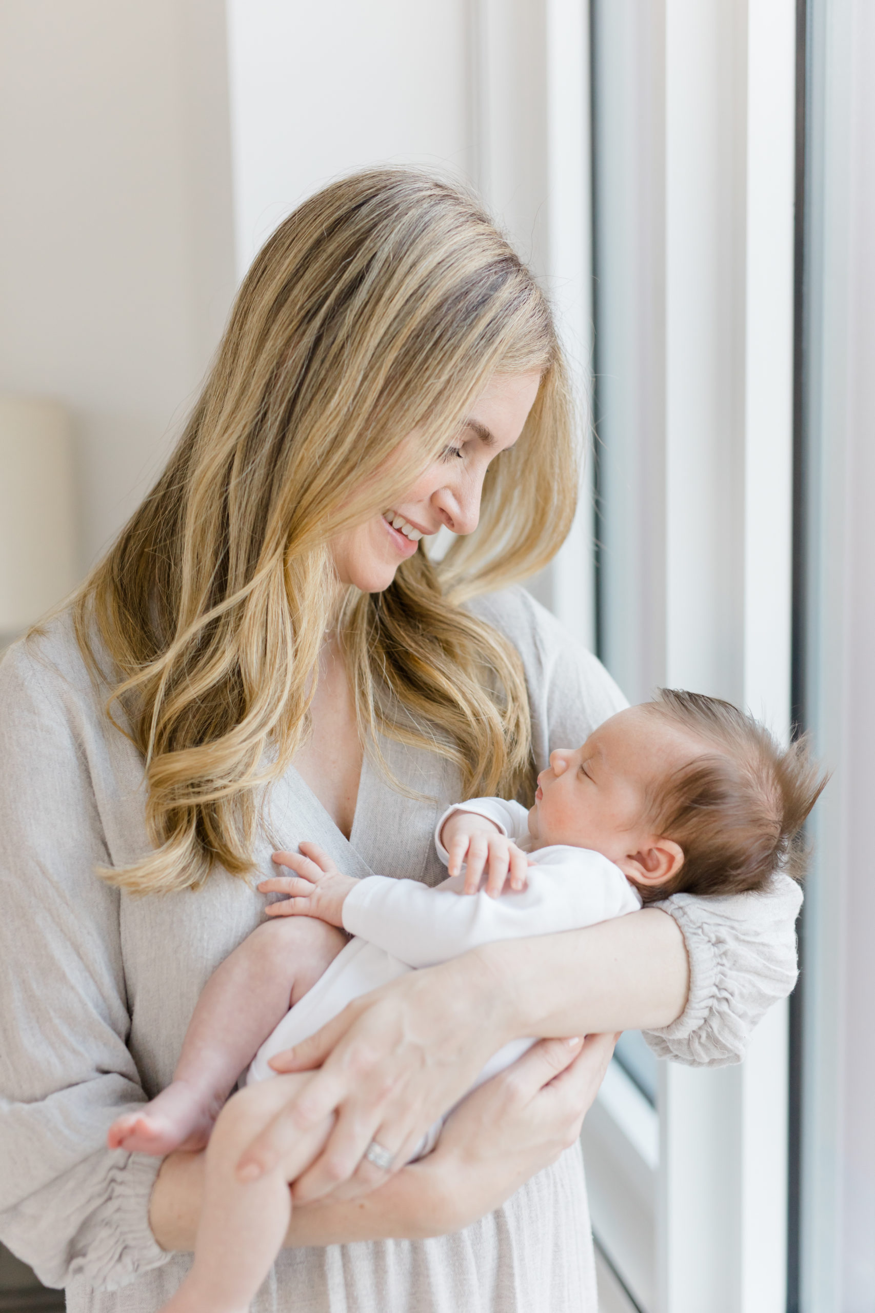 A close-up of a blond mom holding her baby near a window at an in home newborn photography session in NYC photographed by Jacqueline Clair Photography. 