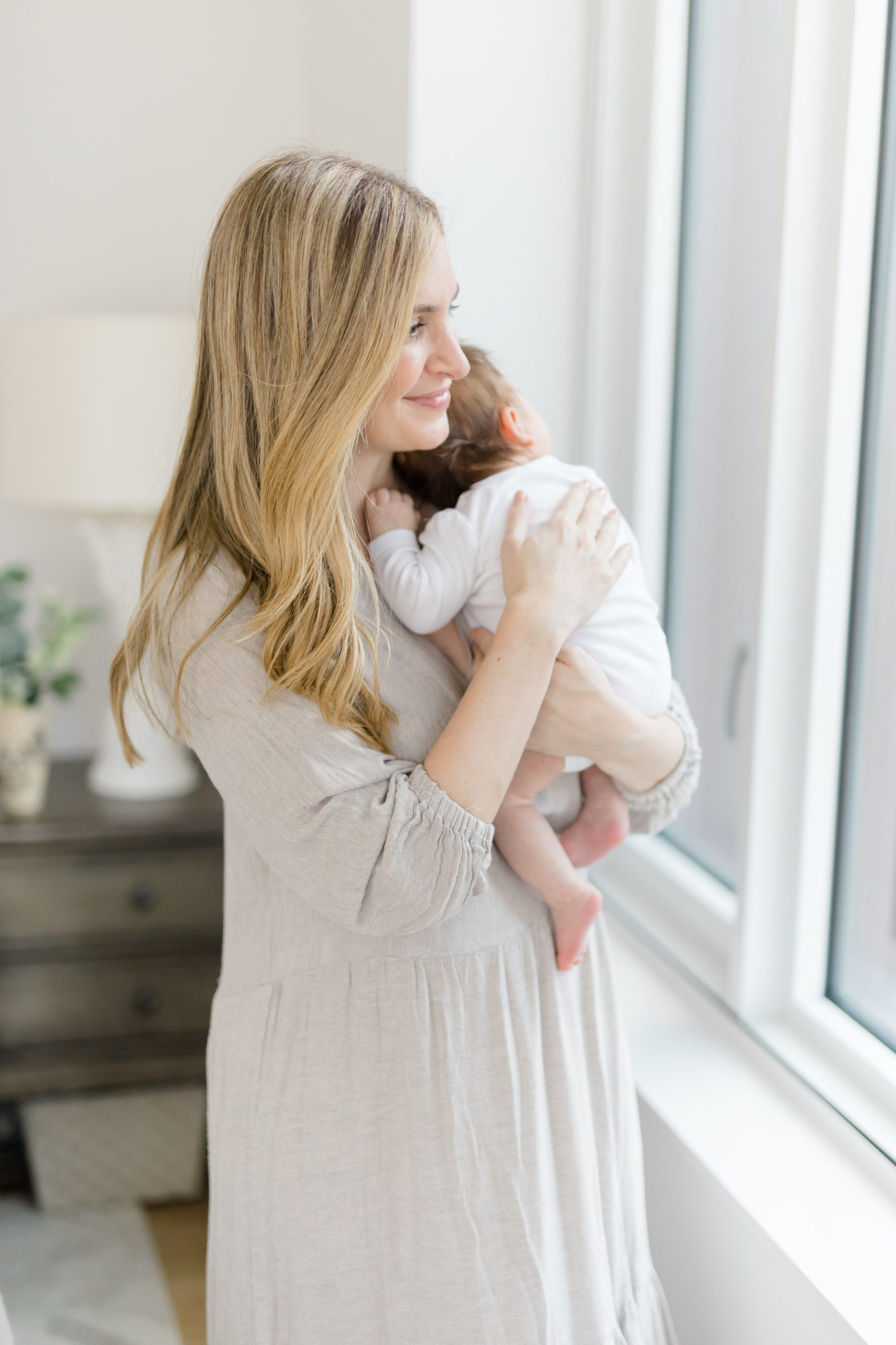 A blond woman holds her baby near a window at an in home newborn photography session in NYC photographed by Jacqueline Clair Photography. 
