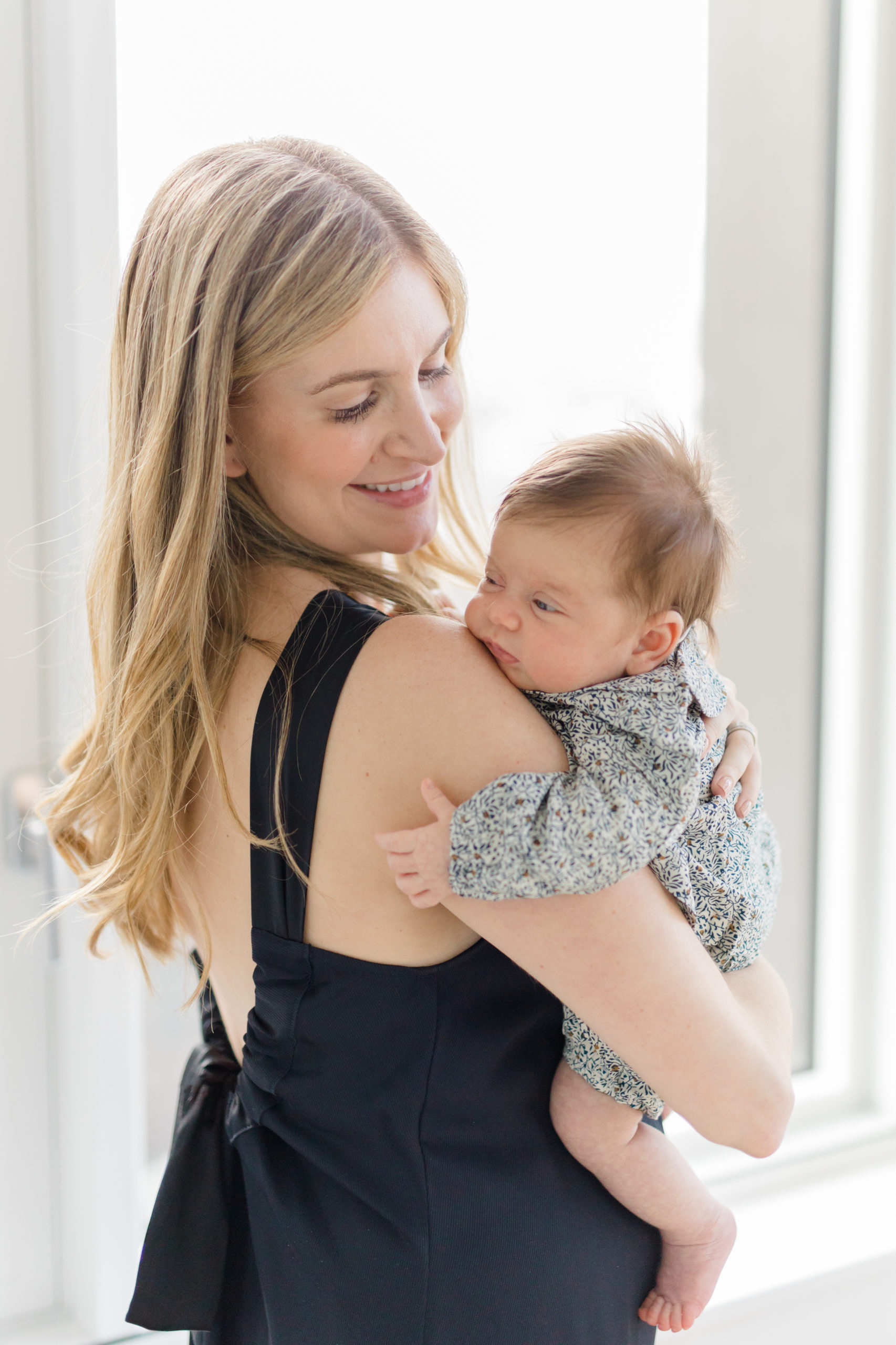 A mom in a black jumpsuit looks over her shoulder at her baby at an in home newborn photography session in NYC photographed by Jacqueline Clair Photography. 
