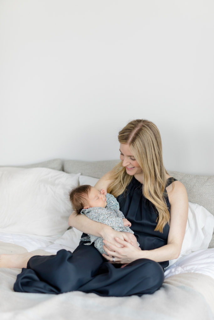 A mom holds her baby while sitting on her bed at an in home newborn photography session in NYC photographed by Jacqueline Clair Photography. 