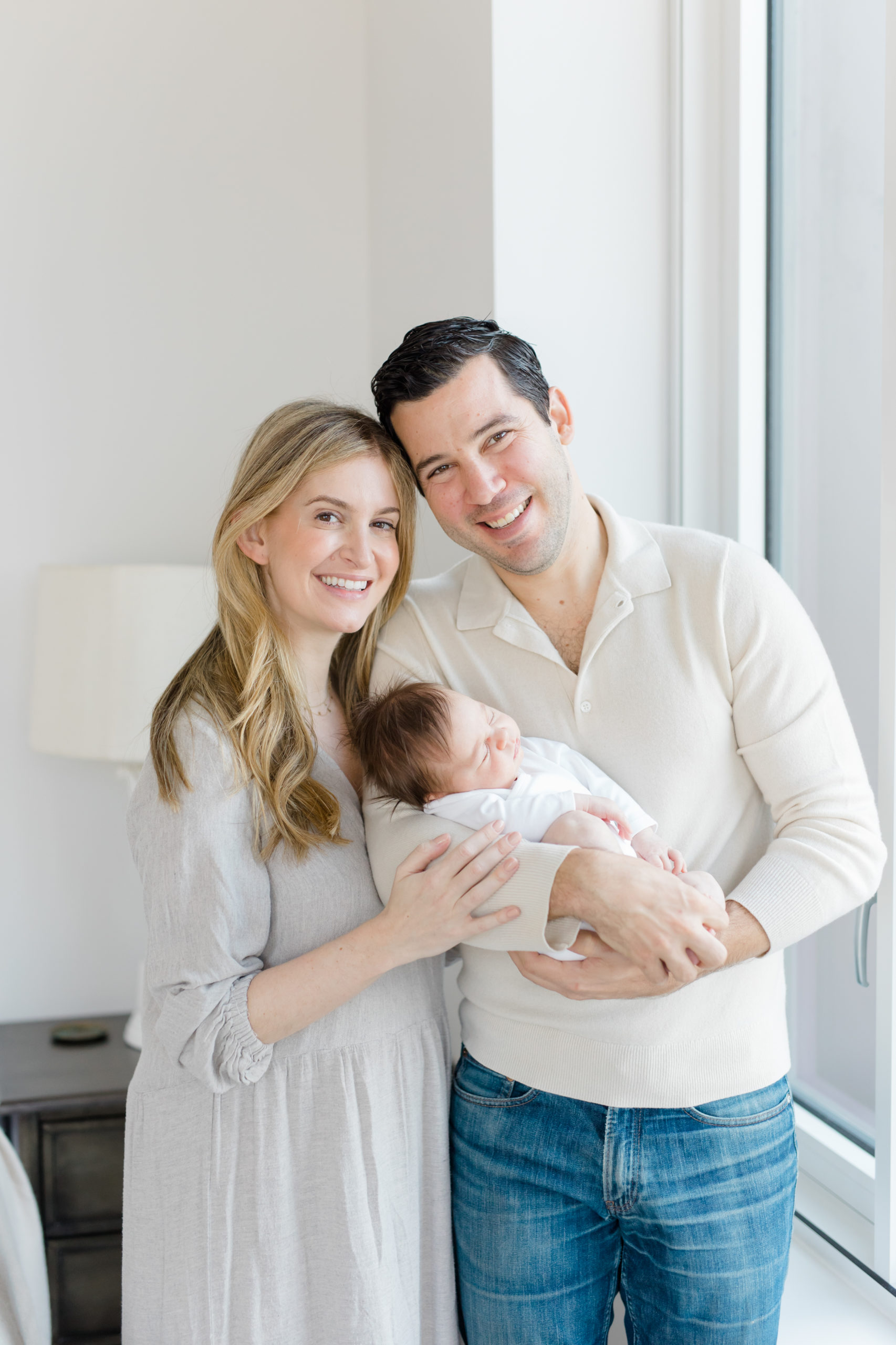 A mom and dad hold their baby near a window at an in home newborn photography session in NYC  photographed by Jacqueline Clair Photography. 