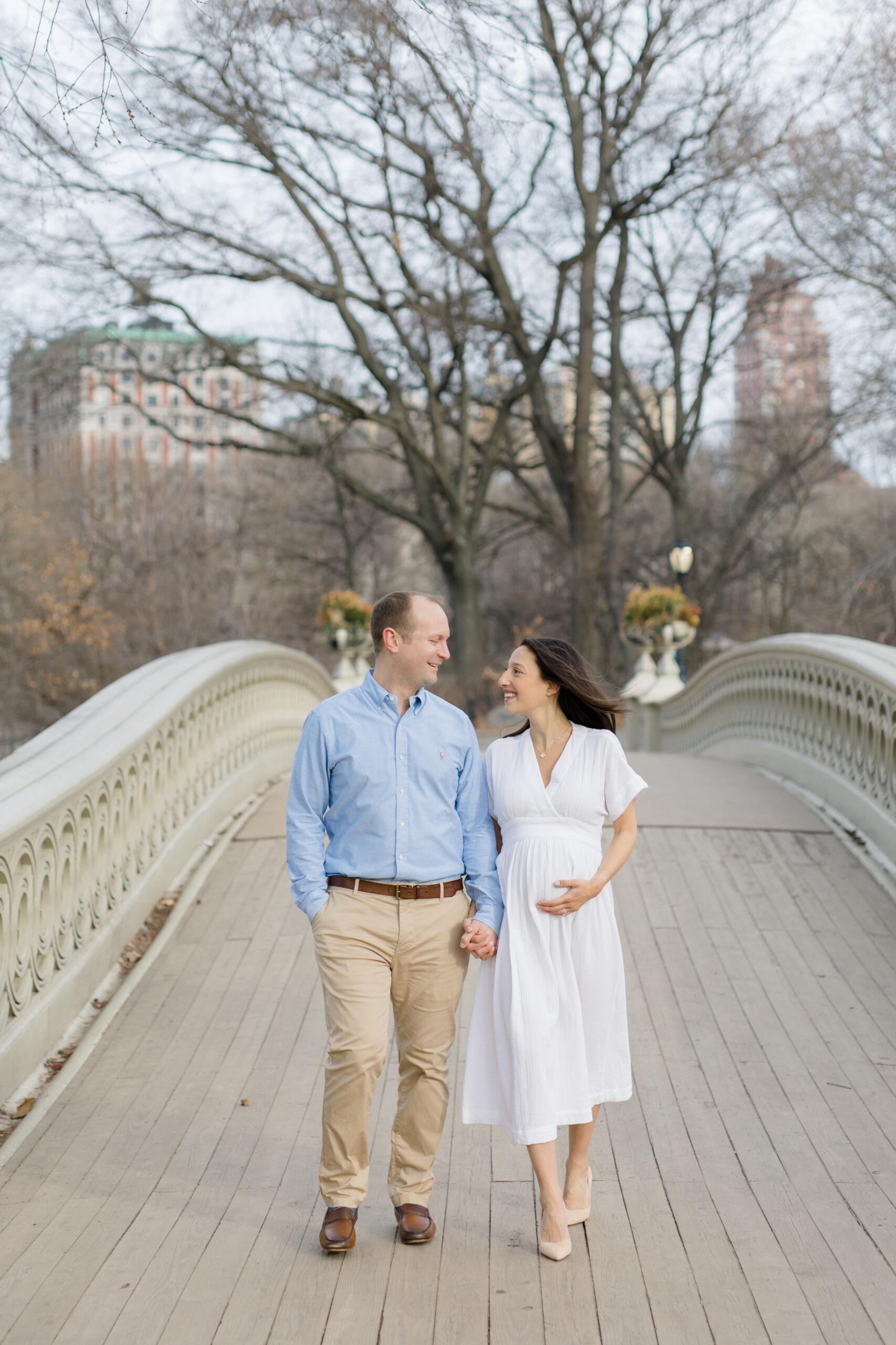An expecting couple walk together on Bow Bridge in Central Park during a New York City maternity photography session with Jacqueline Clair Photography