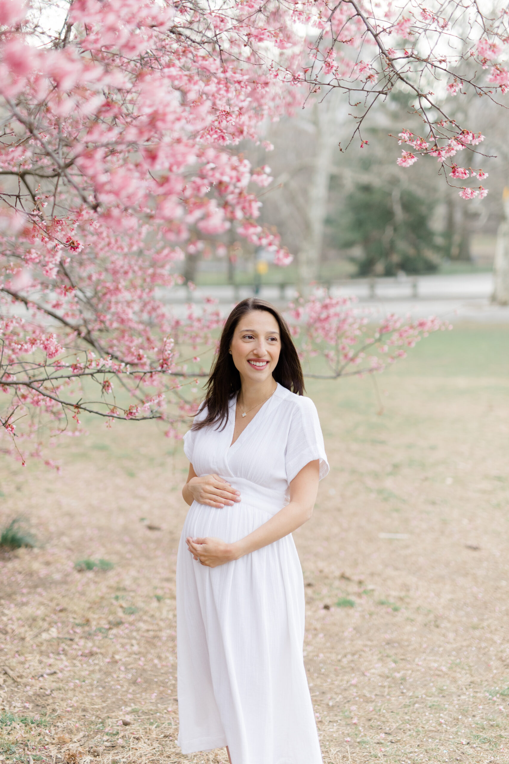 A pregnant woman smiles in front of a pink flowering tree in Central Park at  a New York City maternity photography session with Jacqueline Clair Photography