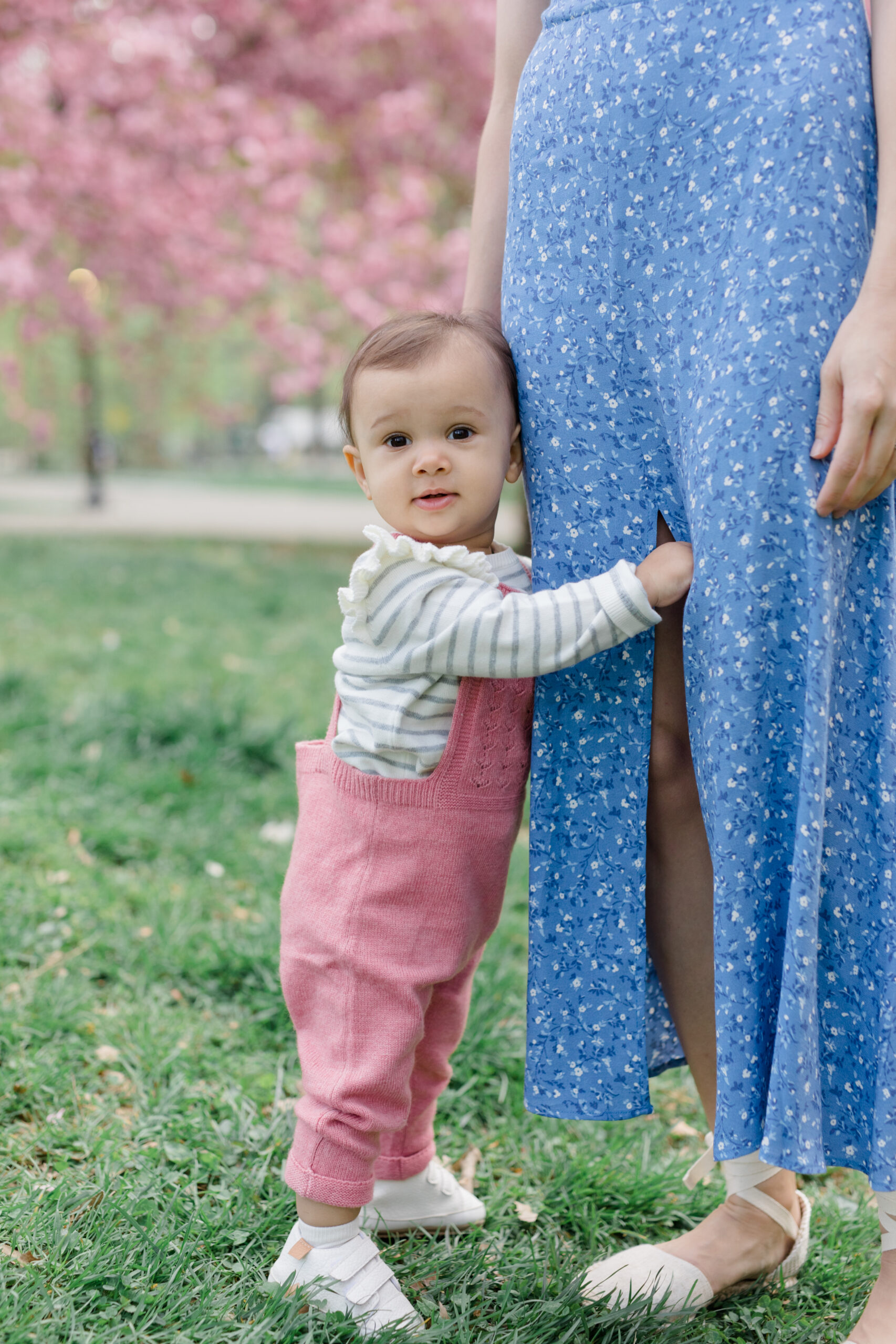 A baby holds her mom's leg at a spring family mini session in Central Park shot by Jacqueline Clair Photography
