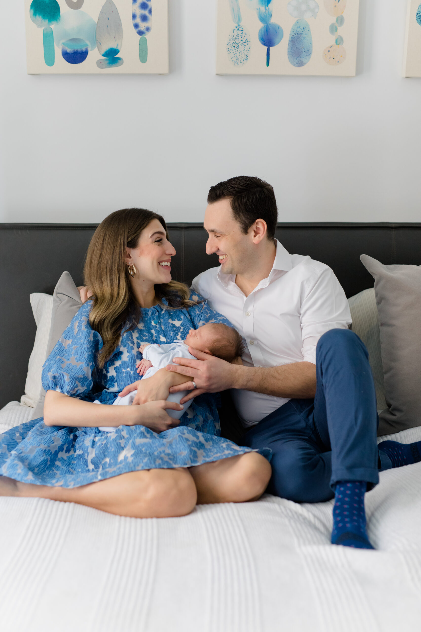 A mom and dad sit on a bed laughing while holding their baby at a newborn session with New York City newborn photographer Jacqueline Clair