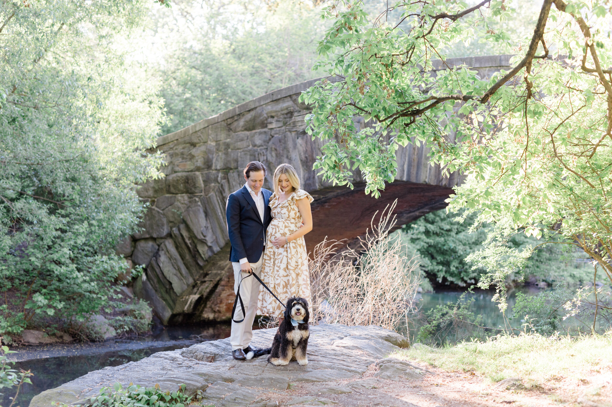 An expecting mom and her husband in front of Gapstow Bridge with their dog at a maternity photo session in Central Park shot by Jacqueline Clair Photography