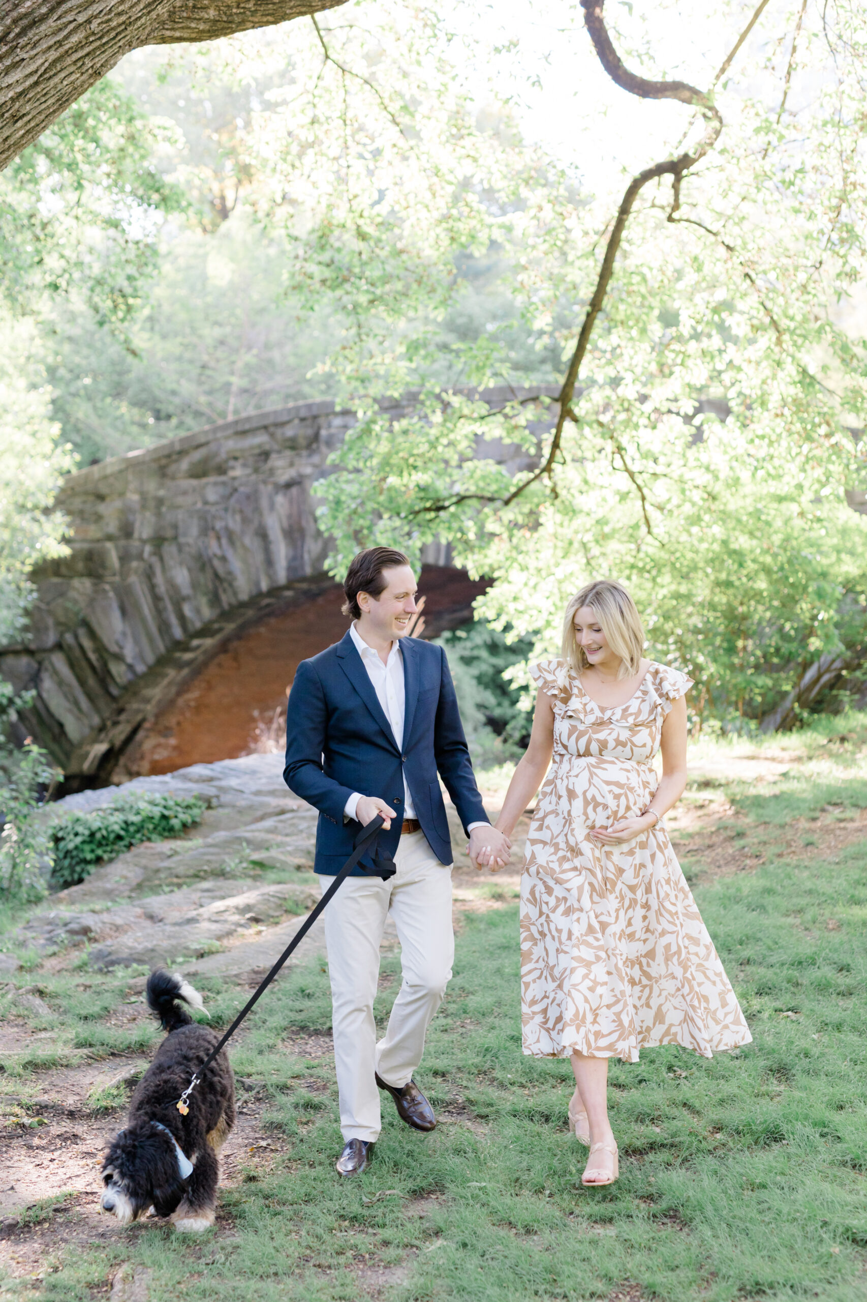 An expecting mom and her husband walk in front of Gapstow Bridge with their dog at a maternity photo session in Central Park shot by Jacqueline Clair Photography