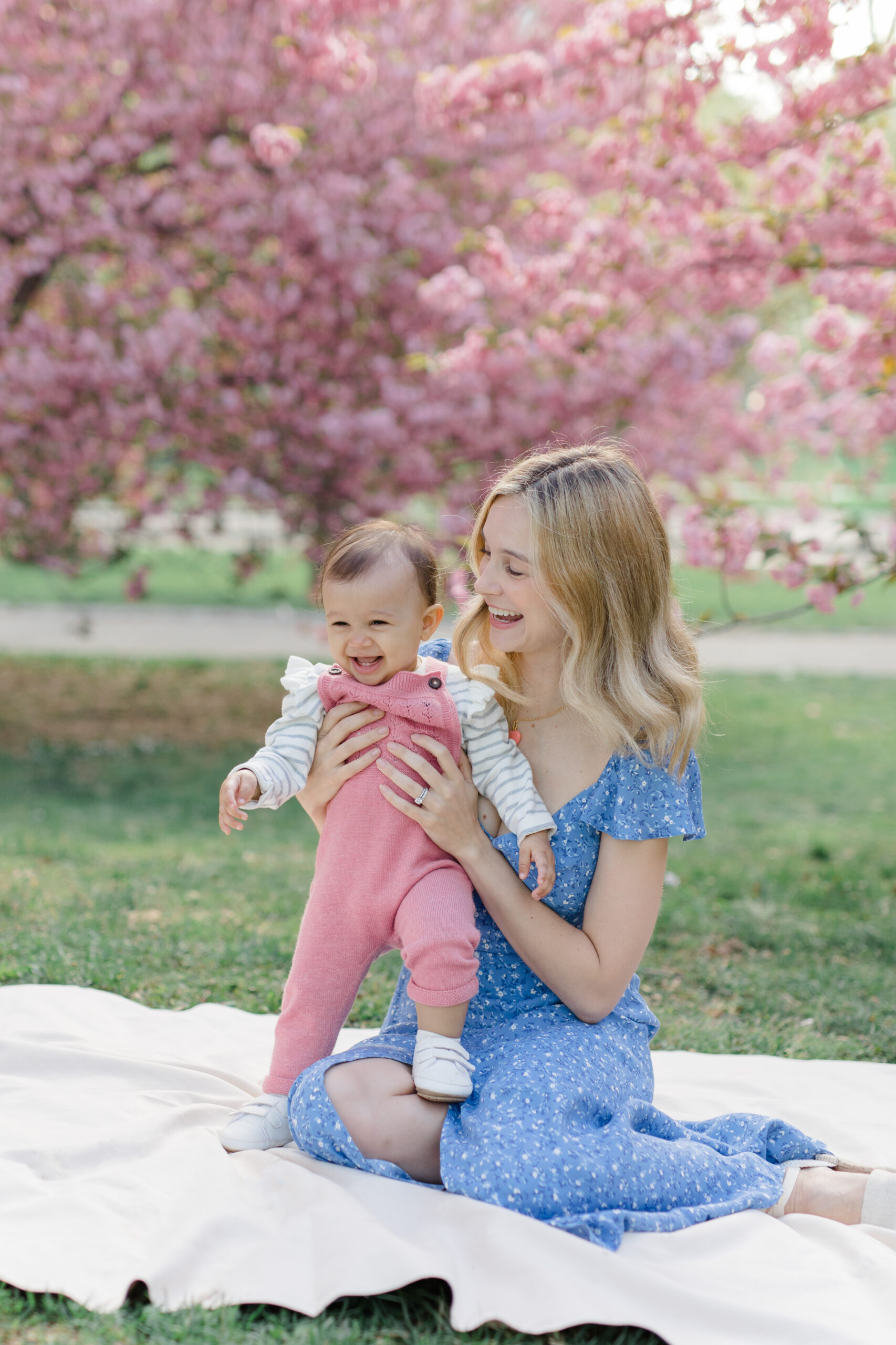 A mom sits laughing with her baby in front of the cherry blossoms at a spring family mini session in Central Park shot by Jacqueline Clair Photography