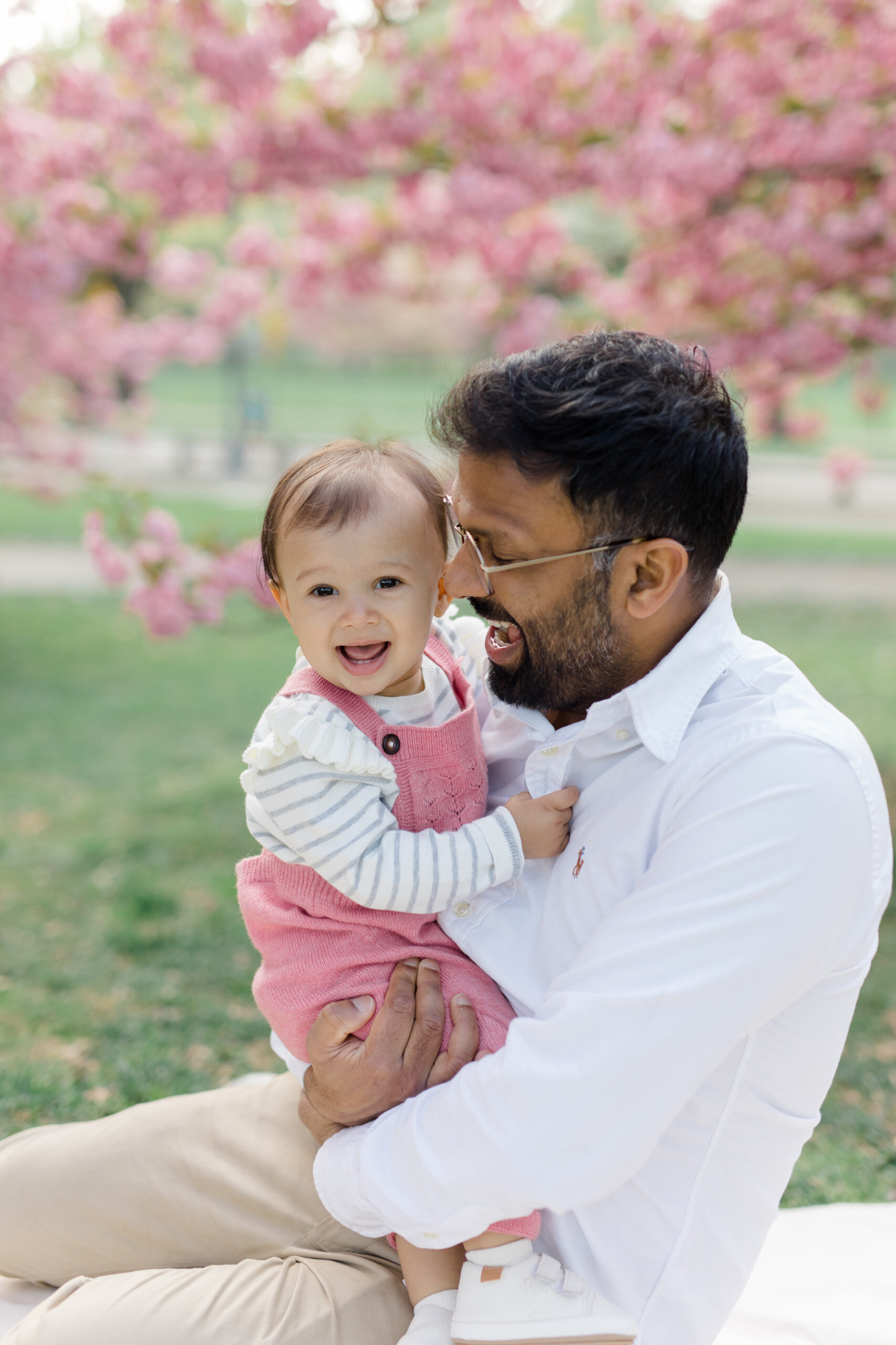 A dad sits holding his baby in front of the cherry blossoms at a spring family mini session in Central Park shot by Jacqueline Clair Photography