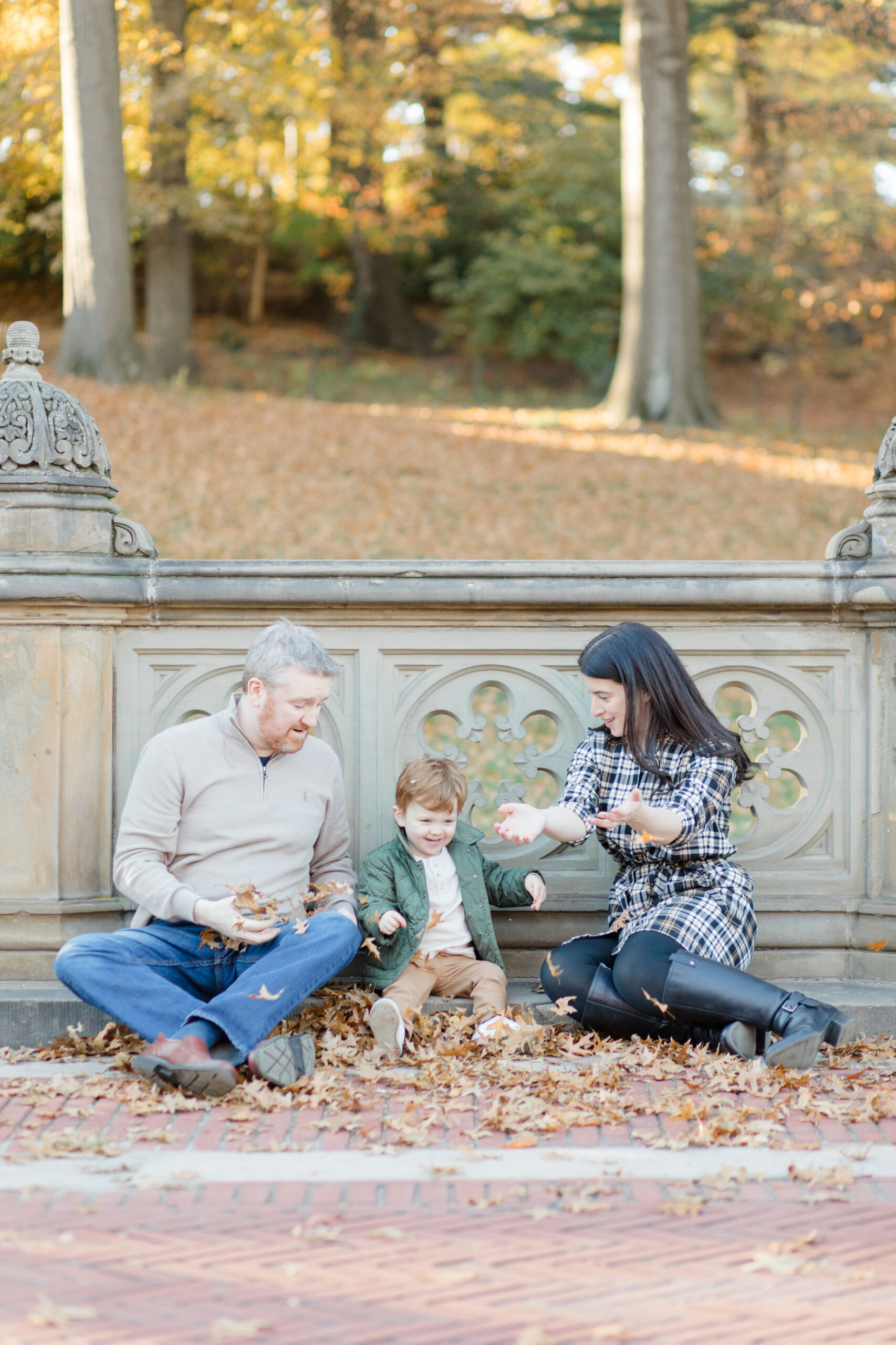 A mom, dad, and their toddler son throw leaves together in Central Park at a fall family session photographed by Jacqueline Clair Photography