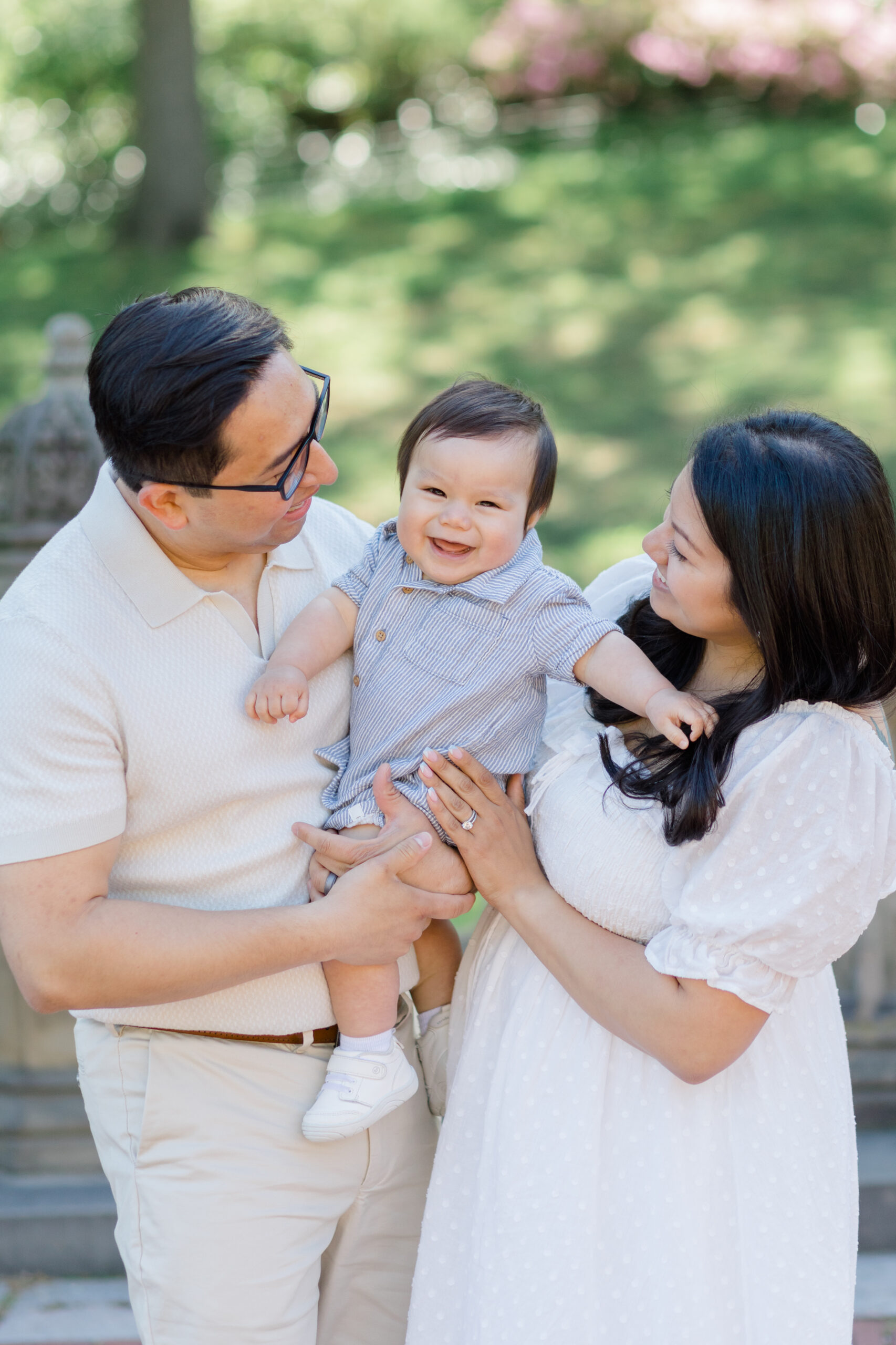 A mom and dad smile at their baby in Central Park, wearing shades of white, khaki, and blue, as an example of what to wear for spring family photos. 