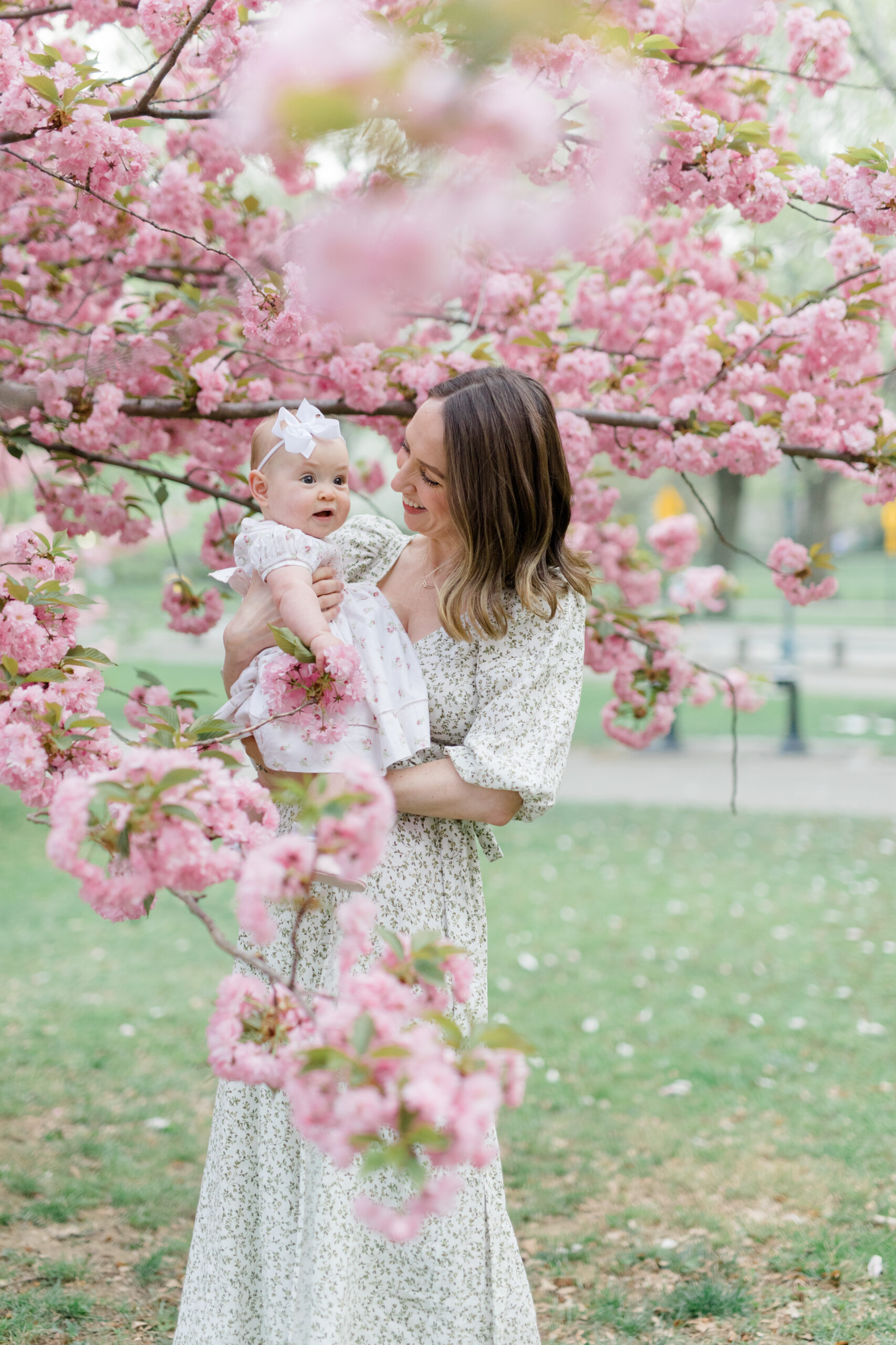 A mom wears a floral patterned dress and holds her baby, also in a floral pattern dress, in front of cherry blossoms, as an example of what to wear for spring family photos. 