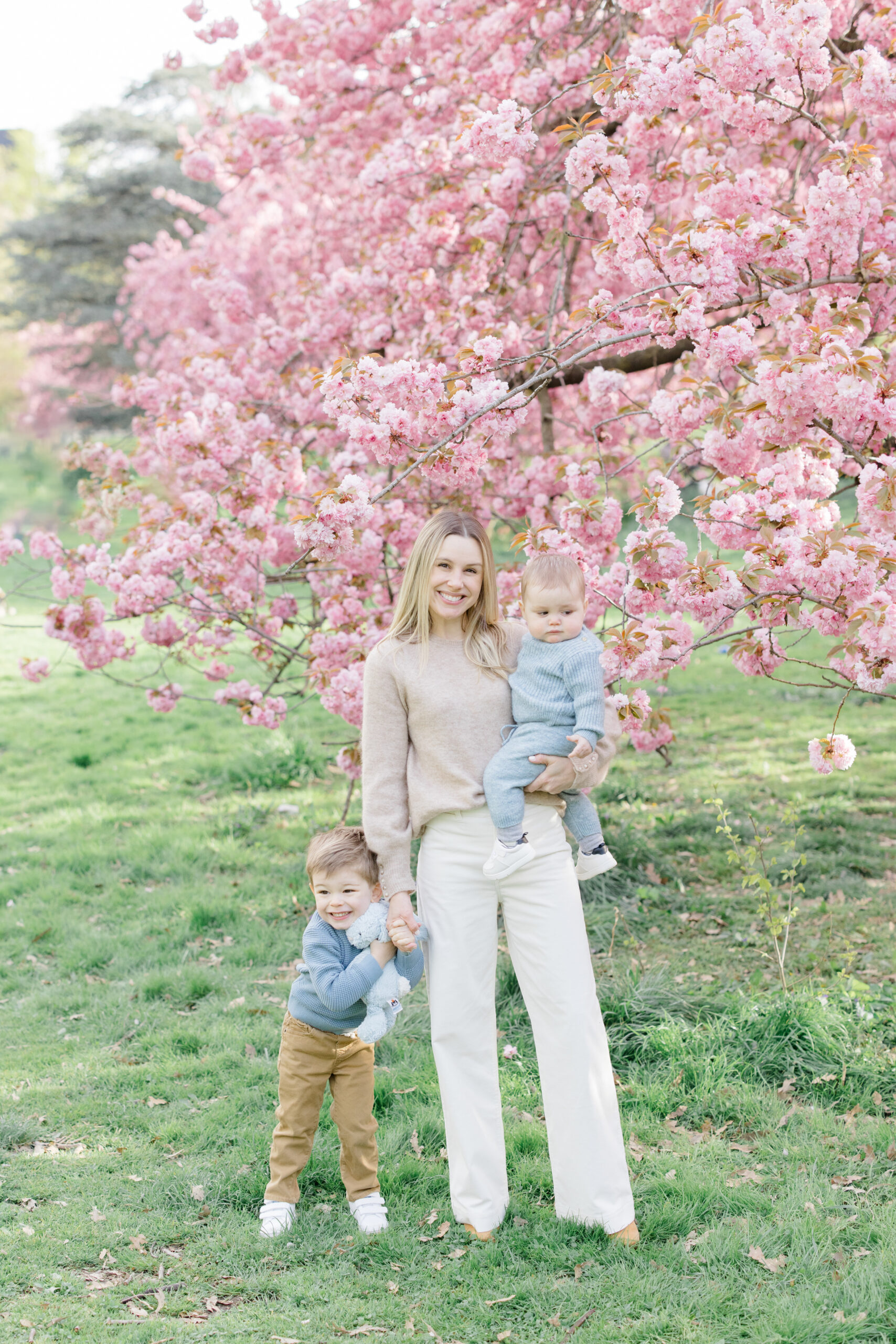 A mom holds her baby son and holds her toddler son's hand as he stands next to her. She is wearing shades of beige and white and the boys are dressed in soft colors of khaki, blue, and gray as an example of what to wear for spring family photos. 