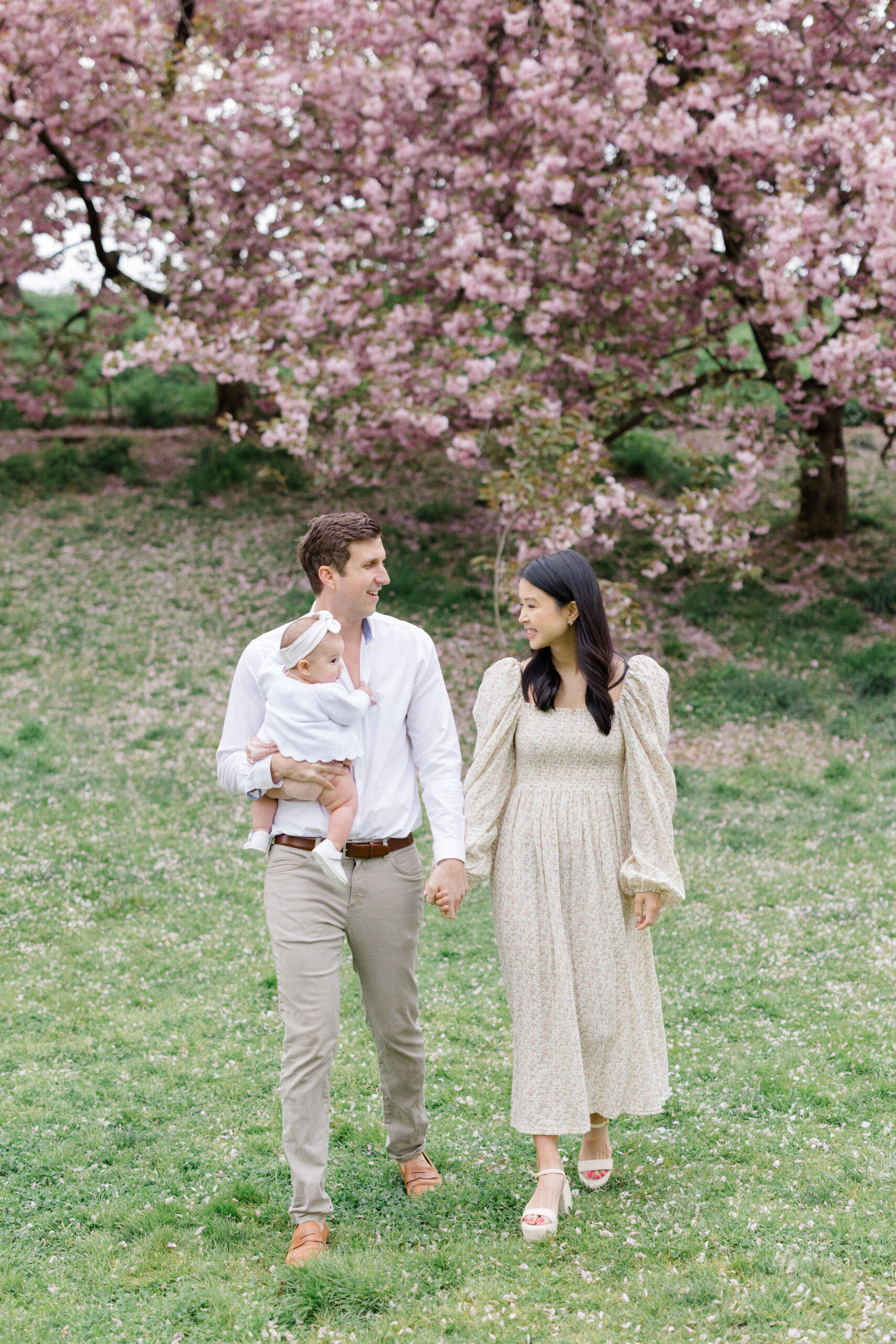 A dad and mom walk with their baby in front of cherry blossoms. Mom wears a long floral patterned dress, dad wears a white button down with khakis, and baby wears a pink set with white sweater as an example of what to wear for spring family photos. 