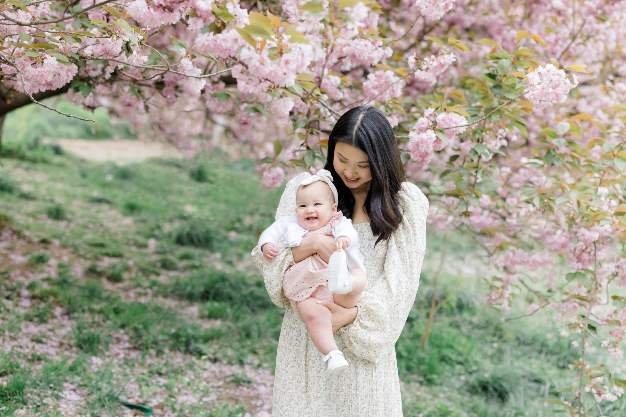 A mom holds her baby in front of cherry blossoms, wearing a softly patterned floral dress with flowy sleeves. Her baby girl wears a light pink eyelet set and a white sweater as an example of what to wear for spring family photos. 