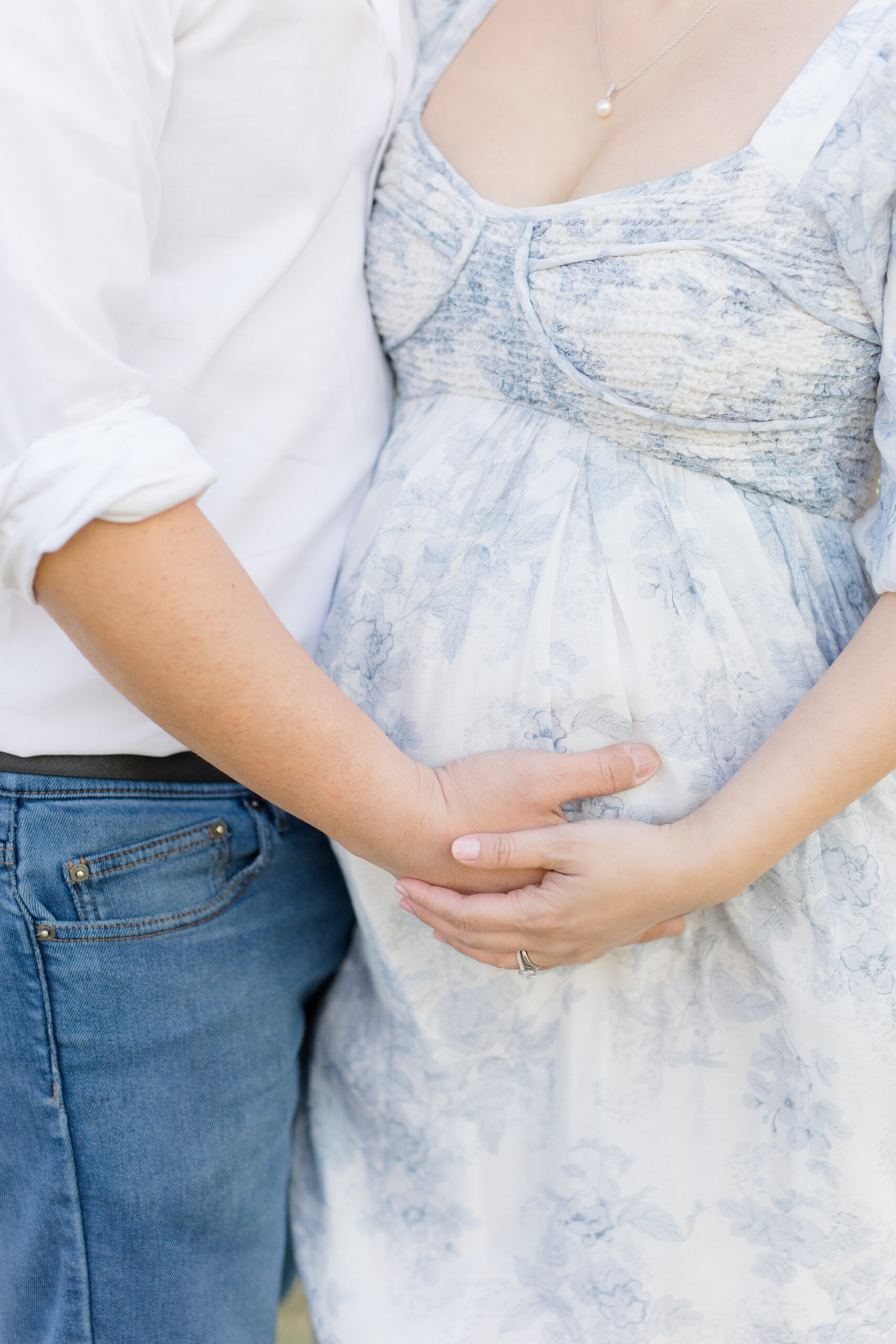 A close up of a pregnant mom's belly with her husband snuggled close. You cannot see their heads, just their torsos, and mom is wearing a softly patterned blue dress as an example of what to wear for spring family photos. 